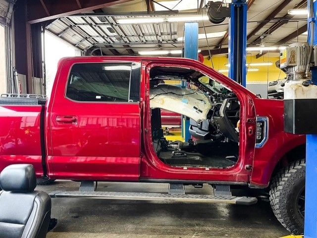 Red pickup truck in a repair shop with its door removed, showing deployed airbags and interior damage.