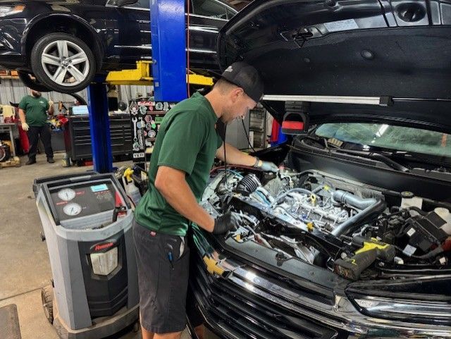 A mechanic works on a car engine with the hood open in a garage. He wears a green shirt and a hat, and there's a machine to his left.