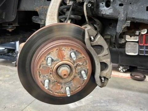 Rusty car brake rotor with caliper and suspension components exposed, in a garage setting.