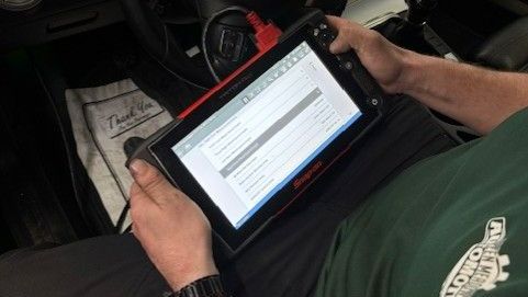 Person in a car, using a diagnostic tablet. Dashboard gauges are lit. Green shirt, black steering wheel.