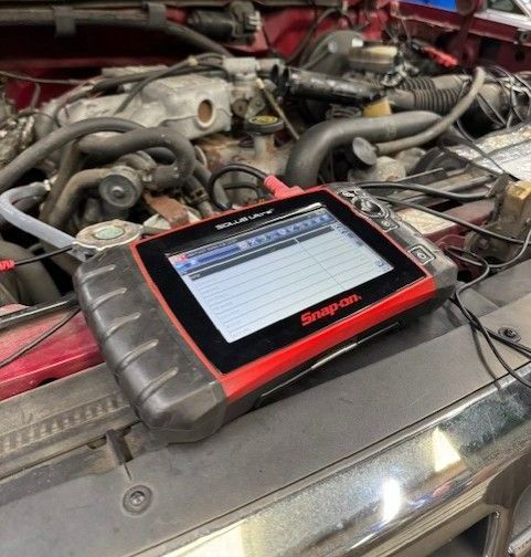 A diagnostic tool, with a display screen showing data, sits on the open hood of a red car. The engine is visible in the background.