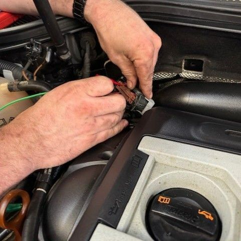 Mechanic's hands connecting wires in a car engine bay, near black plastic components. Focus on the electrical connections.