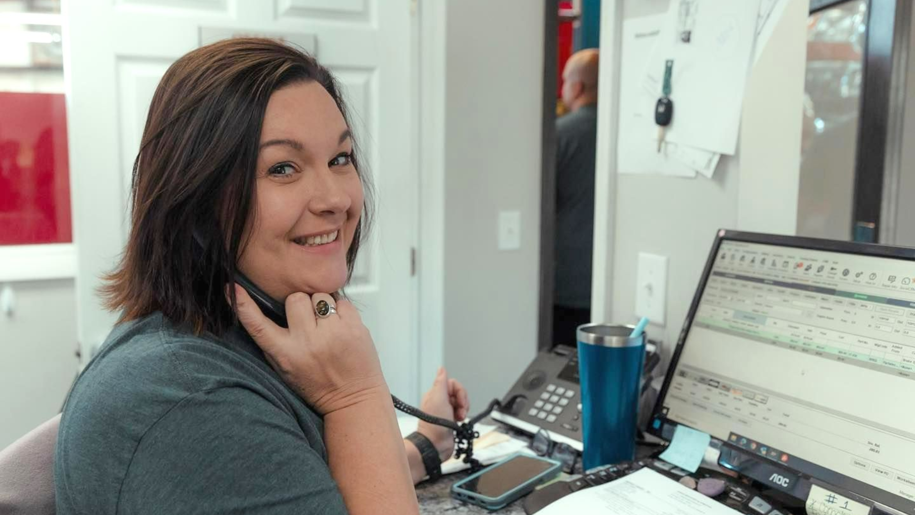 Woman smiling while on the phone at a desk with a computer and papers; a person in the background.
