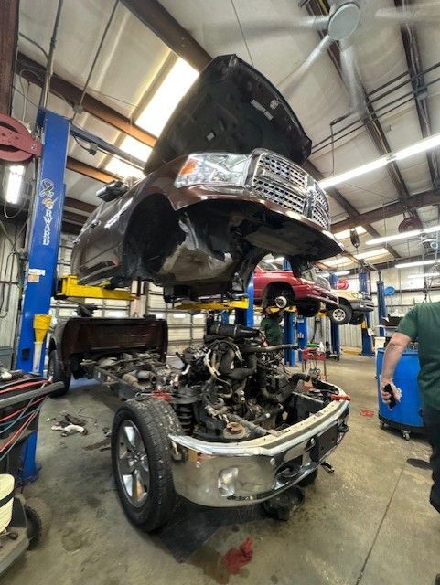 A truck is suspended on a lift in a garage, its body separated from its chassis for repair. A mechanic is nearby.