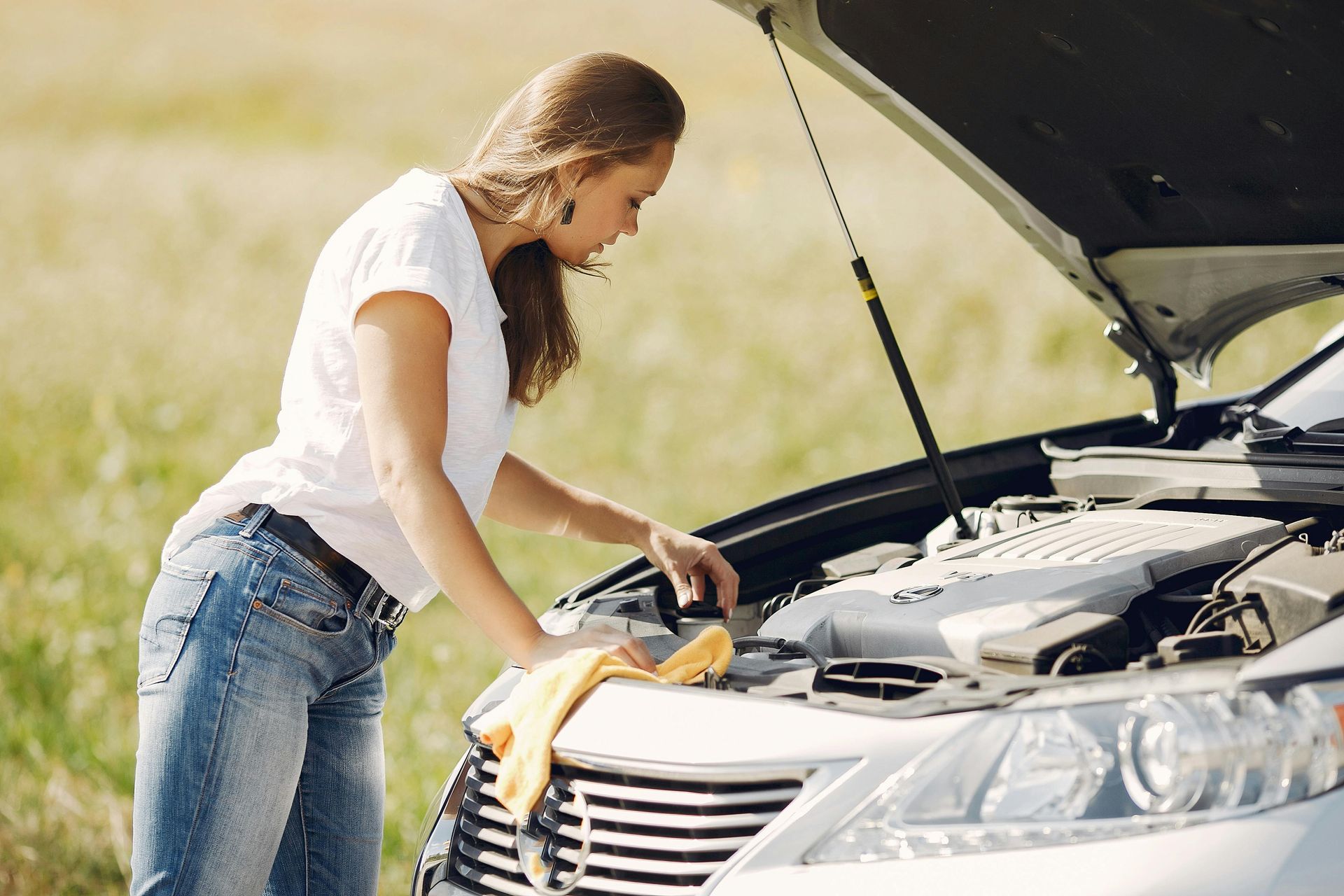 A person in a white shirt and jeans checks the engine of a silver car parked in a field with a yellow cloth in their hand.