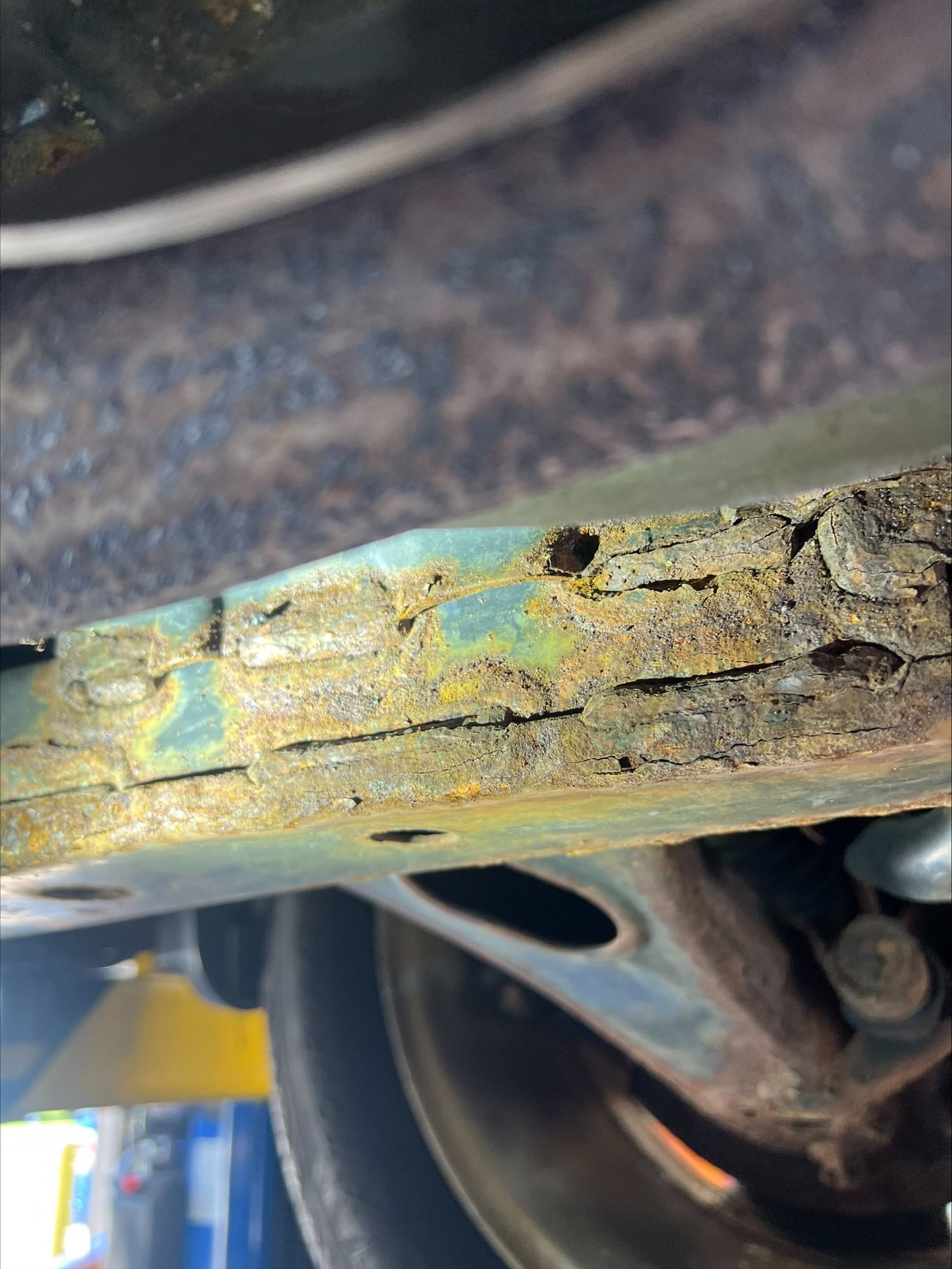 Close-up of a rusty, peeling metal frame with greenish corrosion and a wheel in the background.
