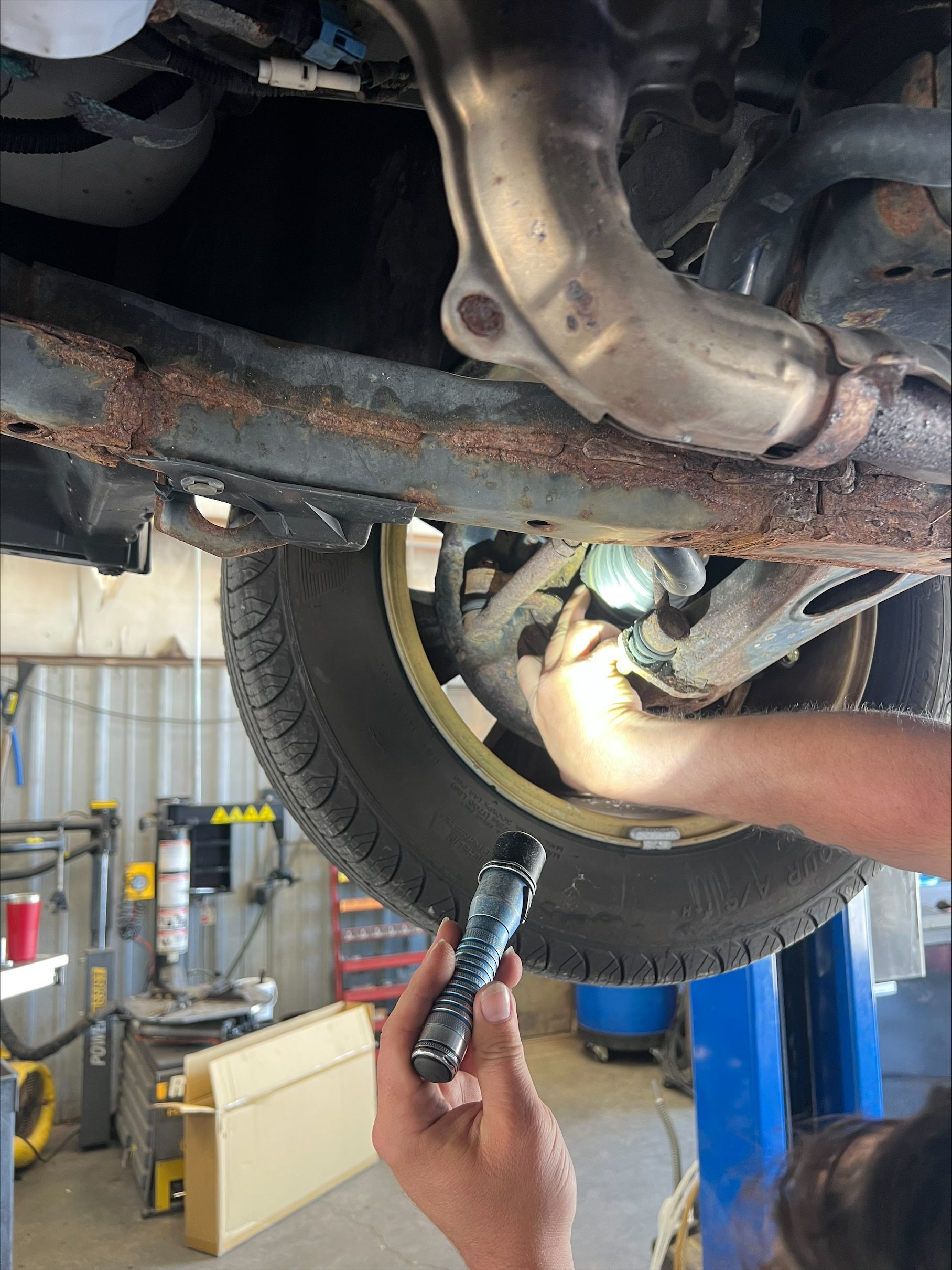 Mechanic inspecting rusted undercarriage of a lifted car with a flashlight in an auto shop