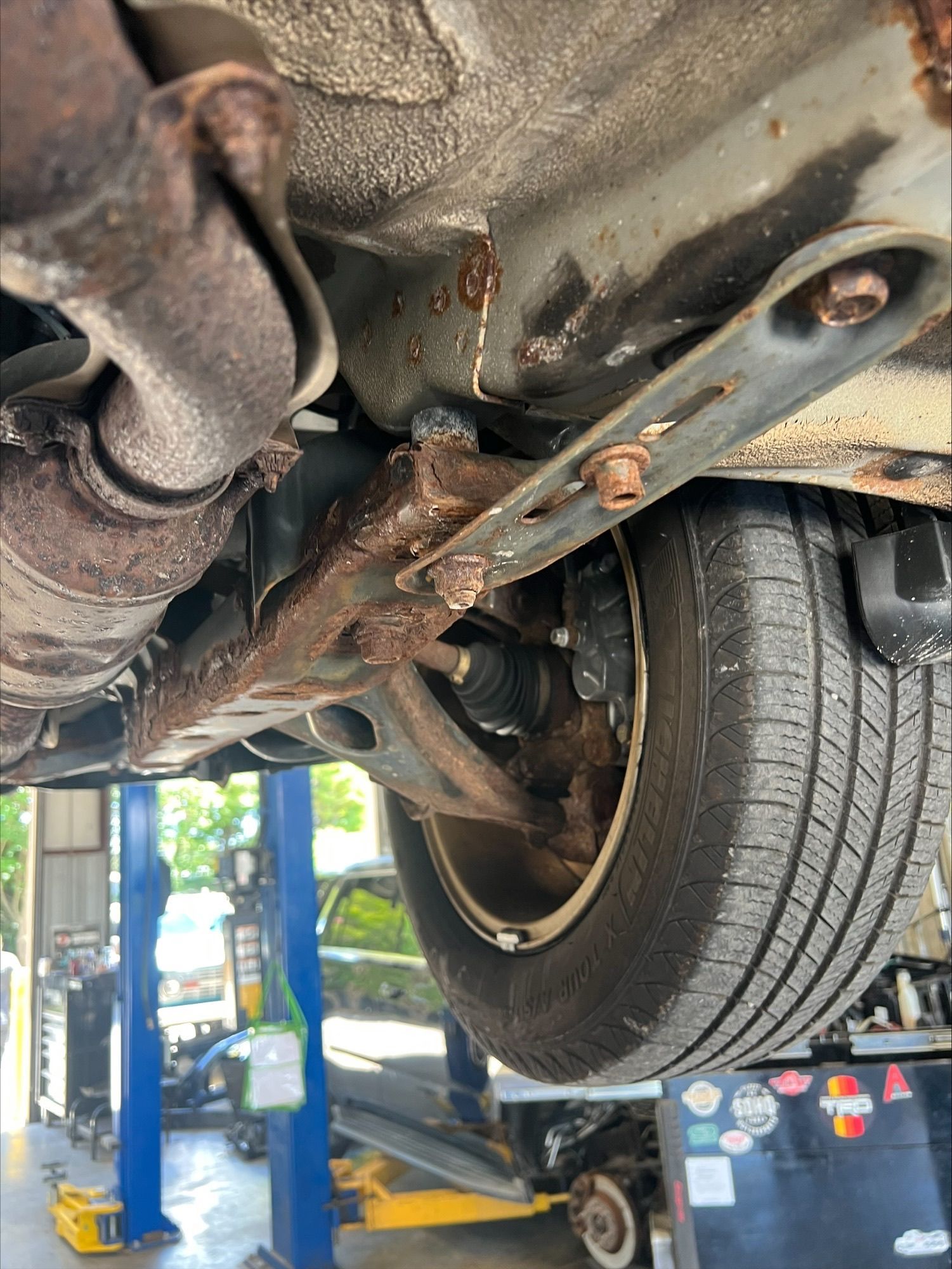 Undercarriage of a car on a lift, showing suspension parts and a rear wheel in an auto repair shop.