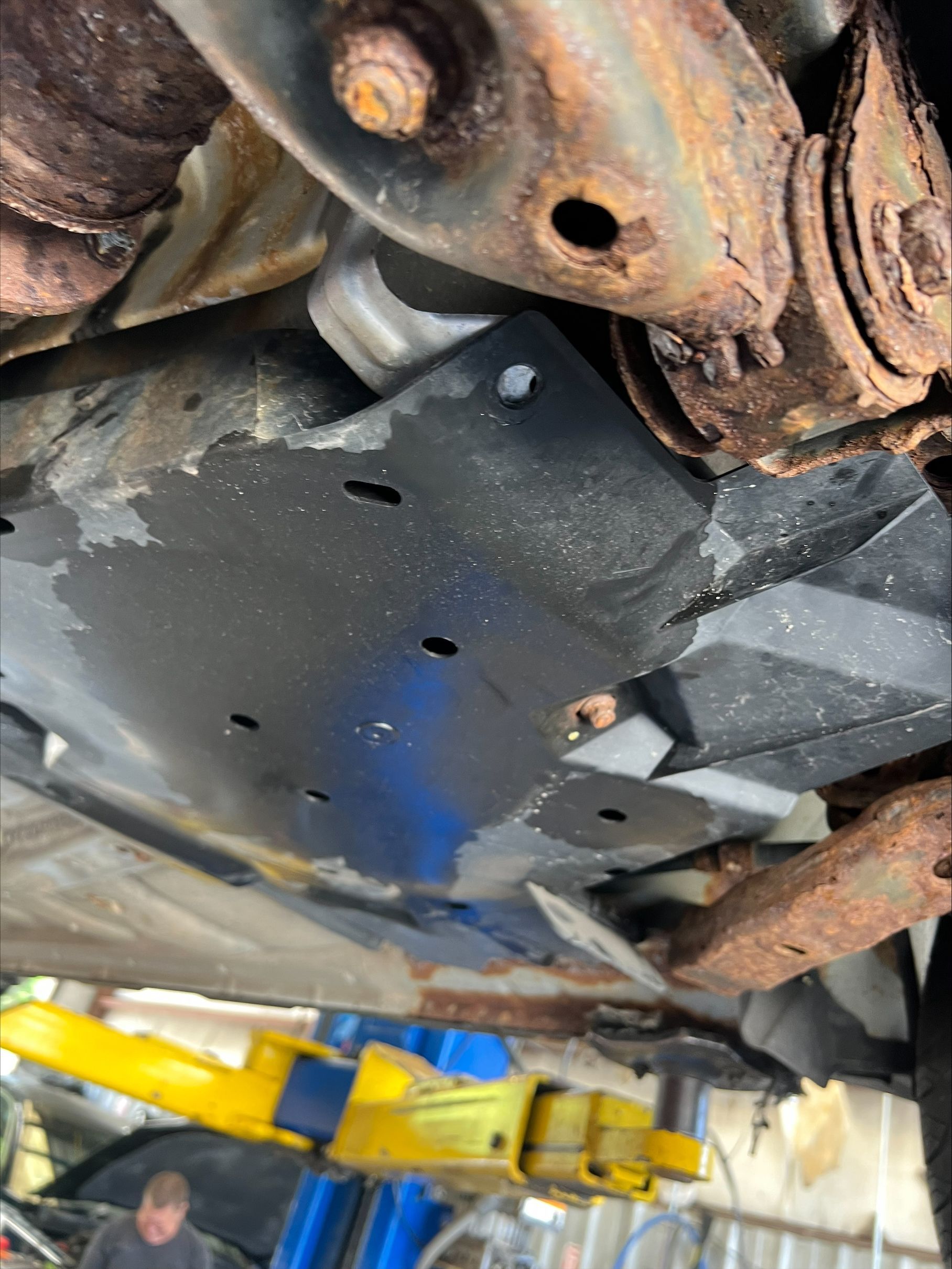 Rusty car undercarriage with a black skid plate and yellow lift arms in a garage