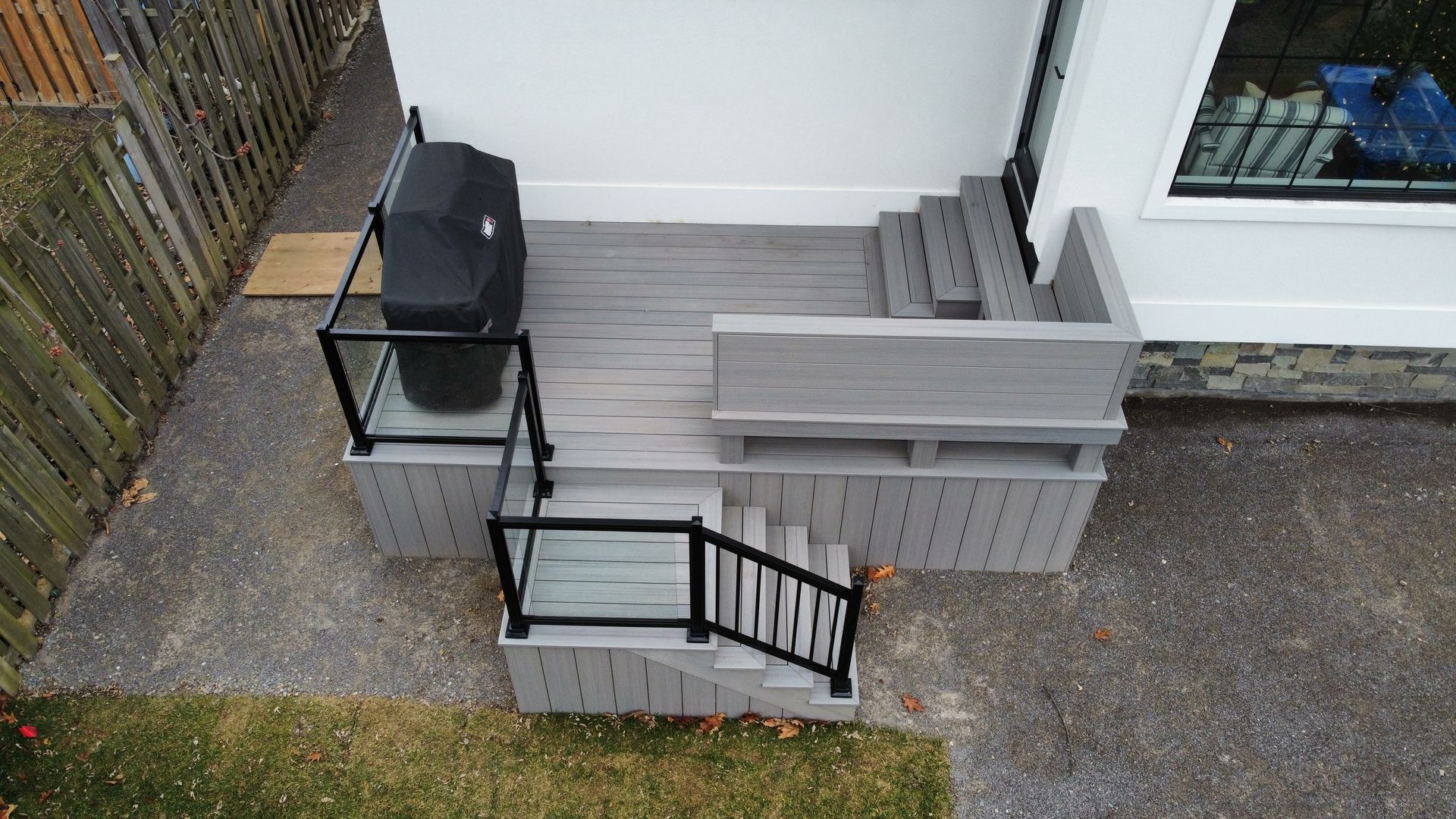 Gray composite deck with a grill, seating, stairs, and glass railings; beside a white building.