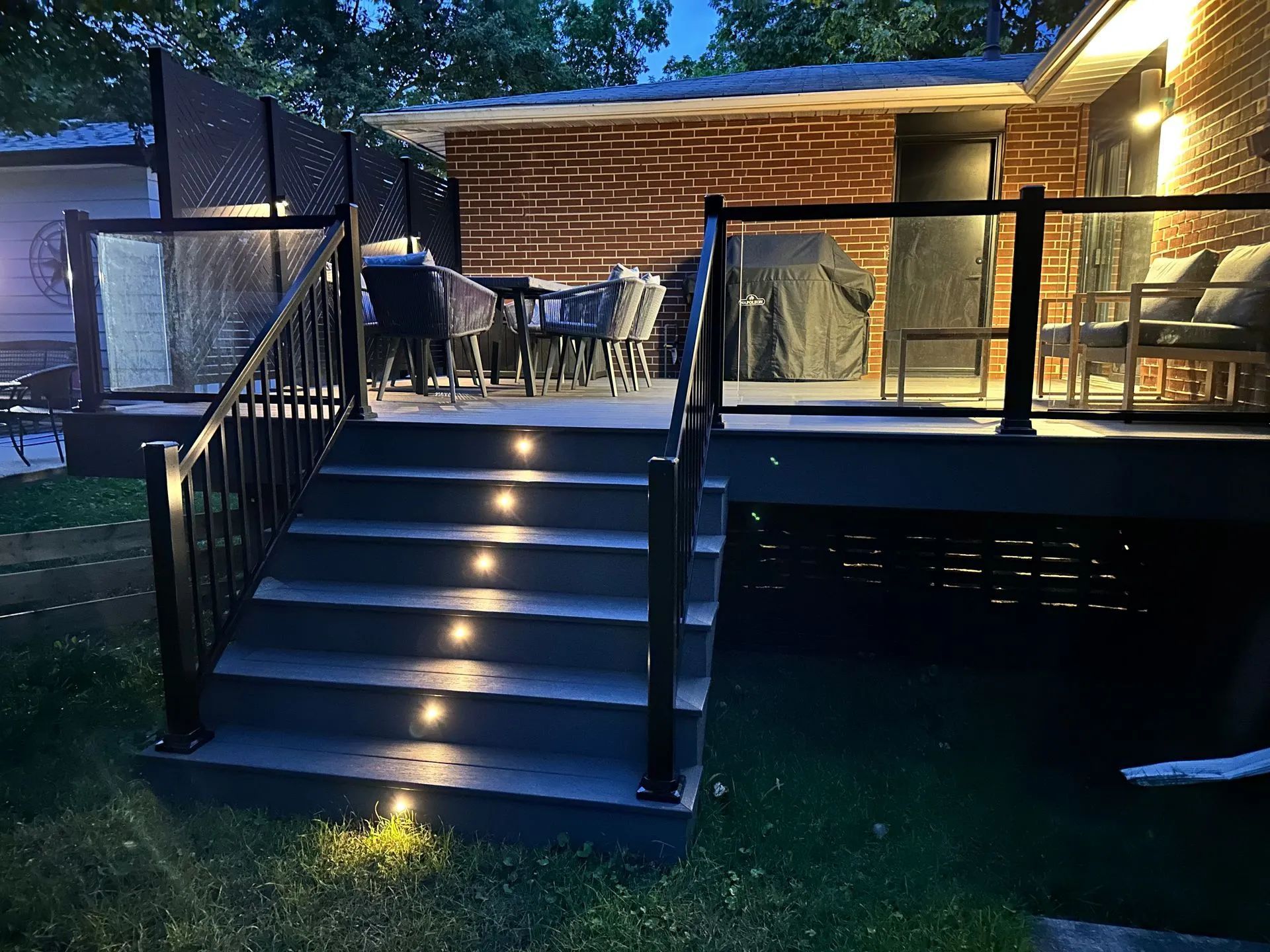 A backyard deck at dusk featuring illuminated stairs leading to a patio area with a dining set and lounge chairs.
