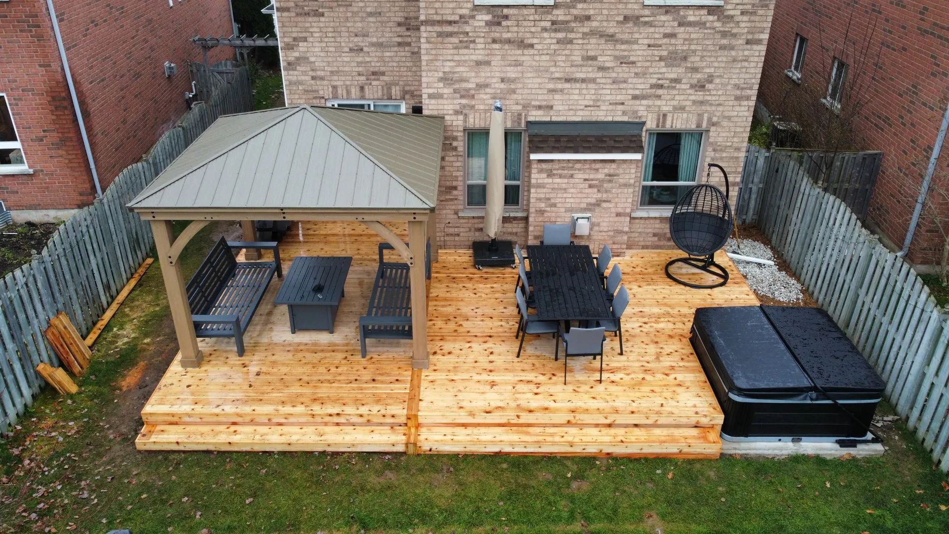 An elevated view of a backyard wood deck with a covered gazebo, dining set, and a hot tub next to a brick house.