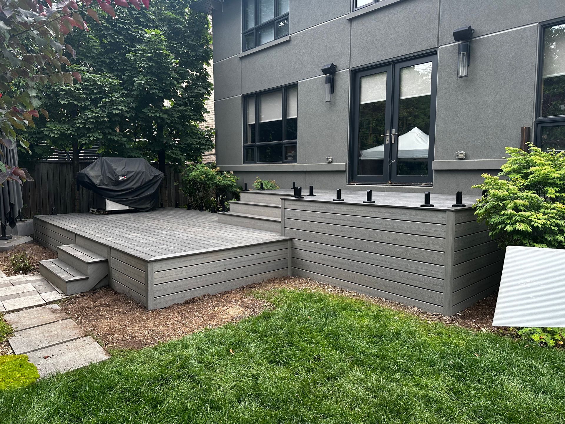 Gray deck and steps lead to a house with dark doors, a grill, and a green yard.