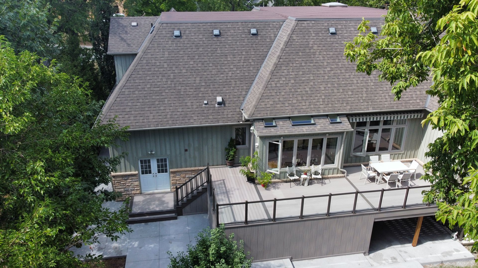 An elevated backyard view of a home with a gray exterior, a large wooden deck, and a stone patio, surrounded by trees.
