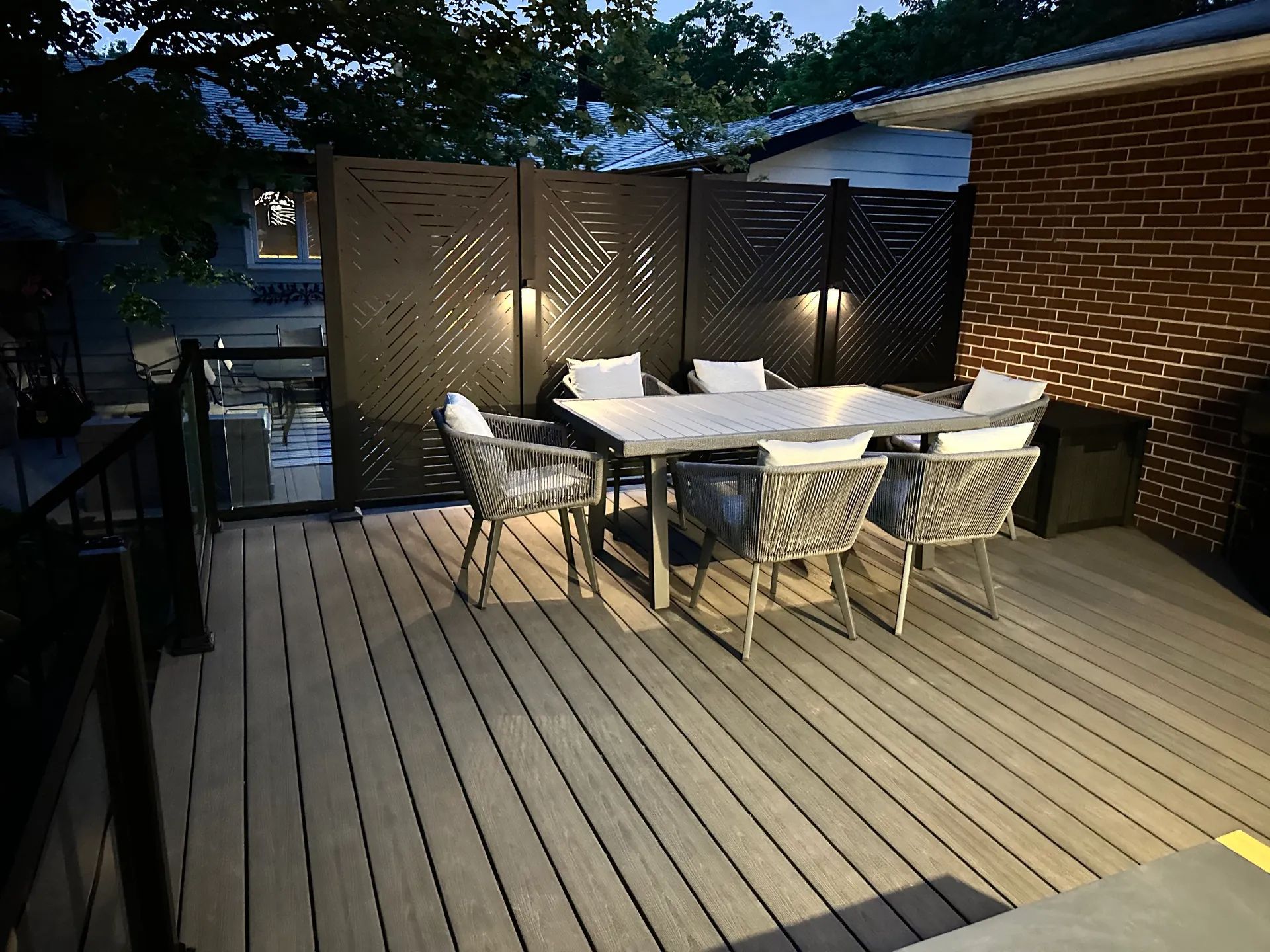 A modern backyard deck at twilight with a patio table, four chairs, and decorative privacy screens illuminated by lights.