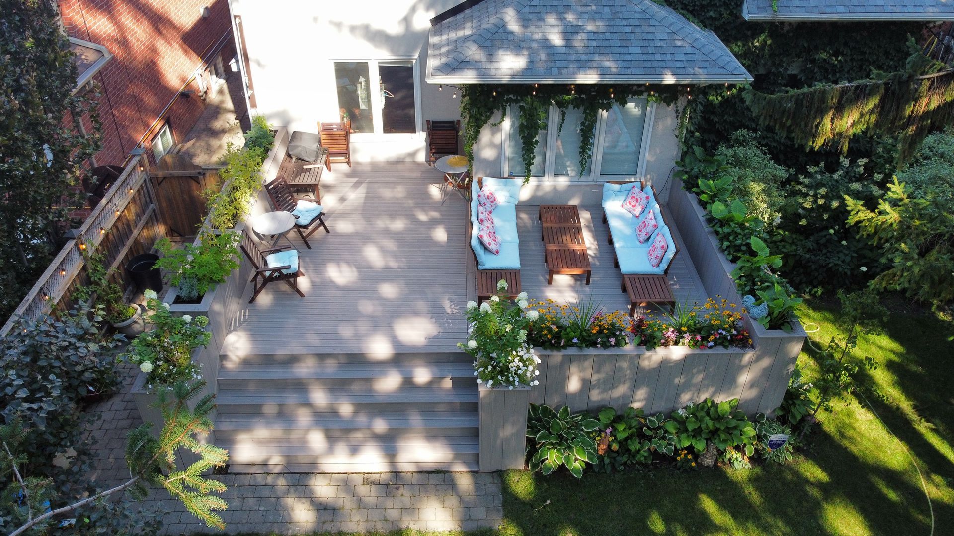 Aerial view of a tiered backyard deck featuring a gazebo with outdoor sofas, a dining area, and lush green landscaping.