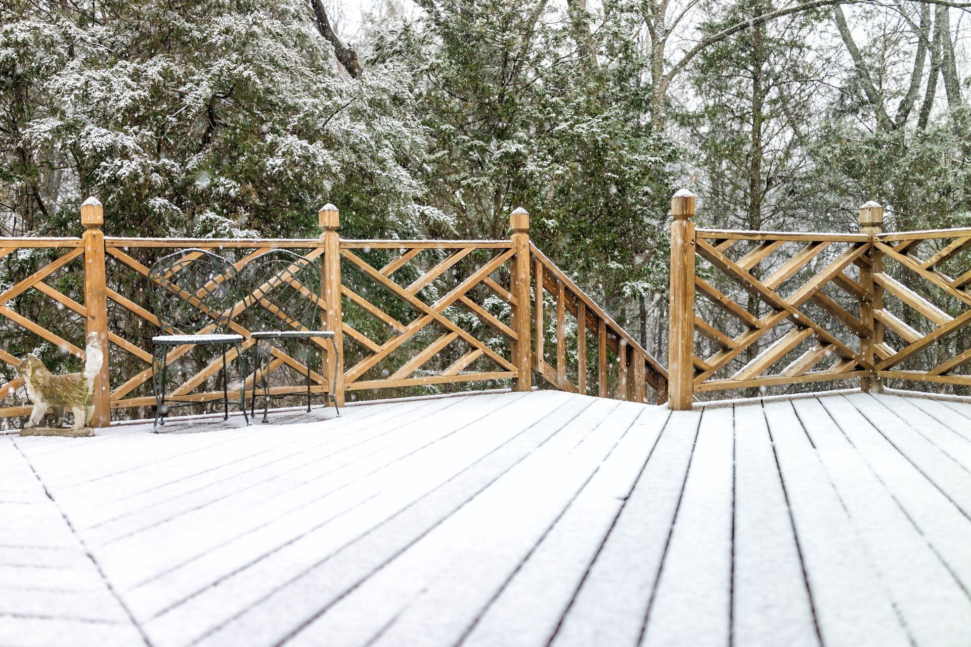 Snow-covered wooden deck with rustic railing, small table, and snowy trees in background.