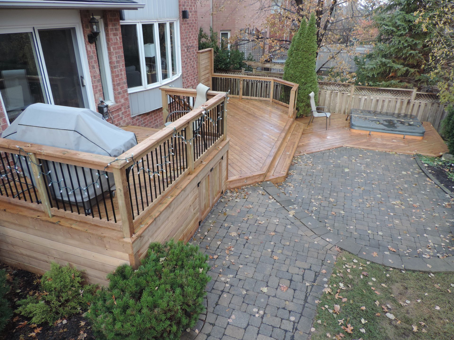 A two-level wooden deck with black railings leads down to a stone patio with a hot tub, set in a backyard.