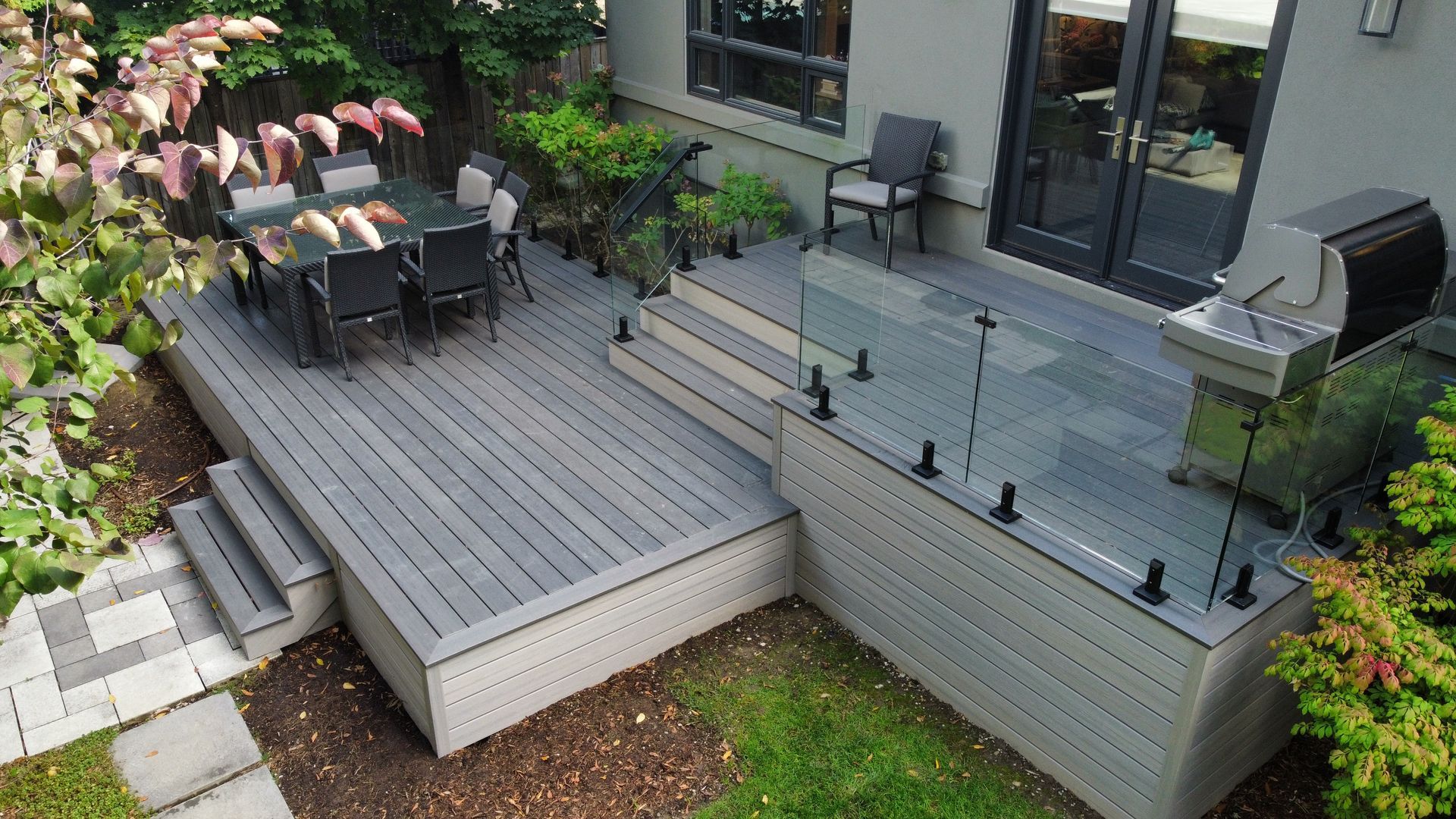 A high-angle view of a grey multi-level outdoor deck featuring a dining area, a grill, and glass railings.