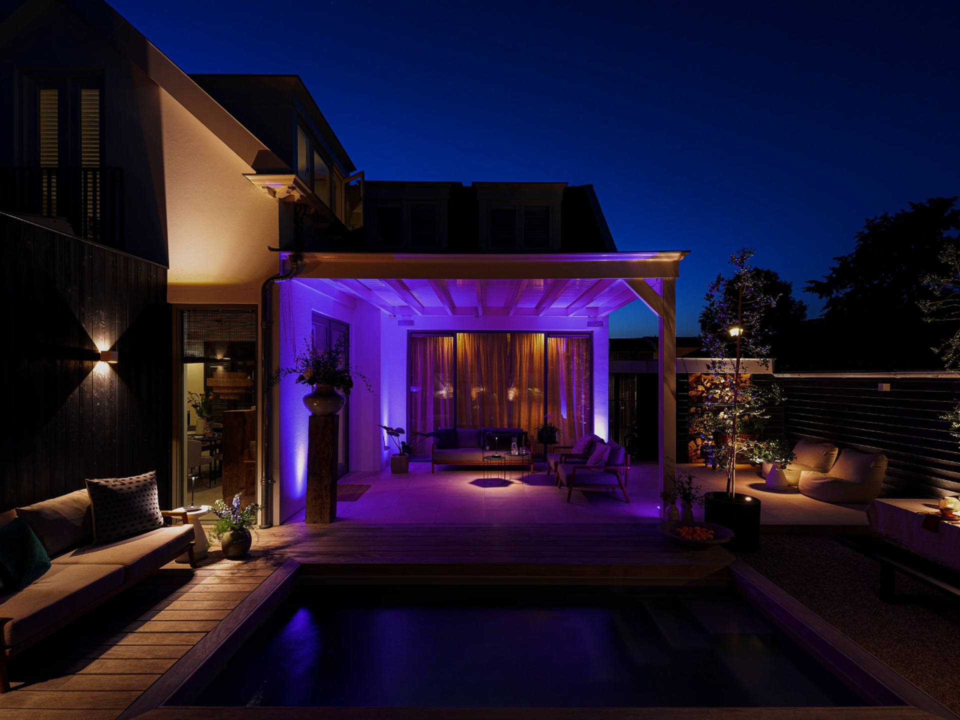 Backyard at night lit with purple lights. Pool, seating, and house.