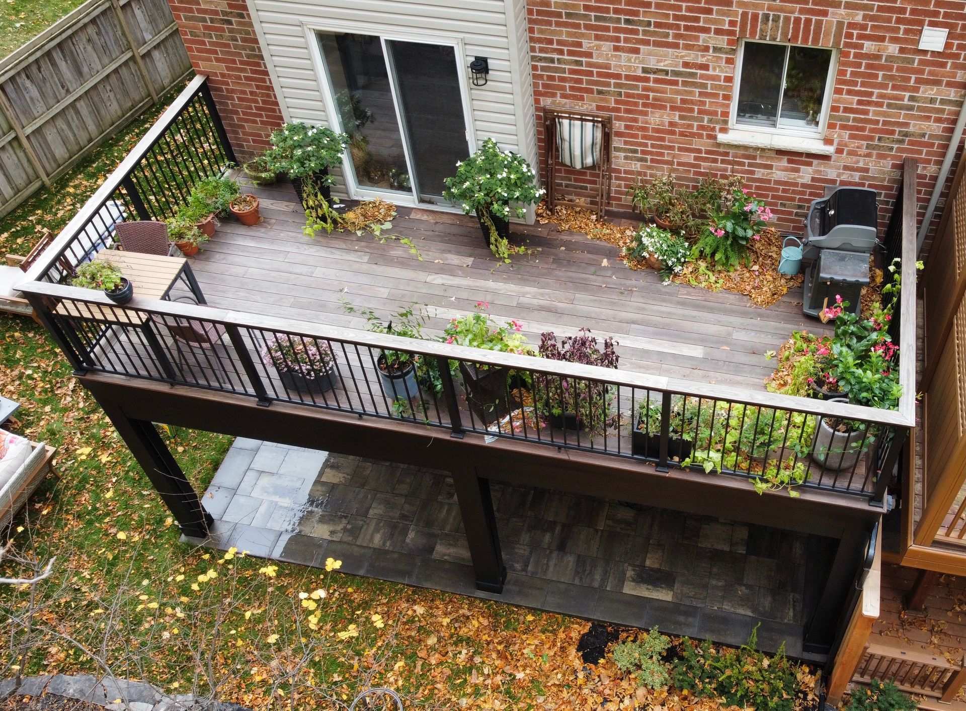 Elevated wooden deck with railing, potted plants, and seating, attached to a brick house. Overlooking a yard with fallen leaves.