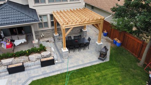 Wooden pergola over a dining table on a patio, adjacent to a house. Green grass and a wooden fence surround it.