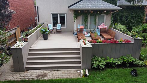 A backyard patio with tiered seating, planters, and a small building with seating, surrounded by greenery.