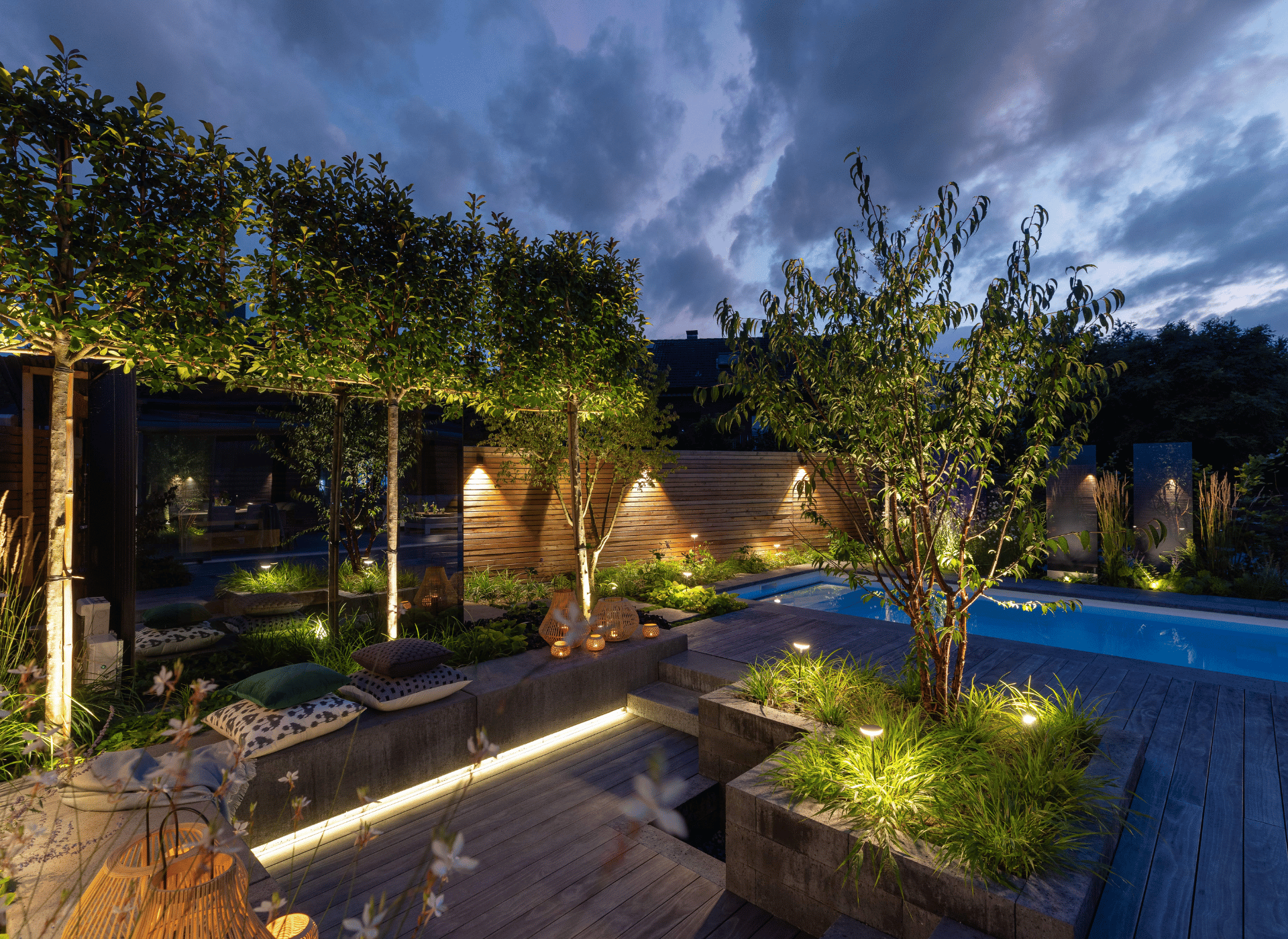 Night-lit garden with trees, a wooden fence, and a pool. Yellow lights illuminate plants, creating a calm atmosphere.