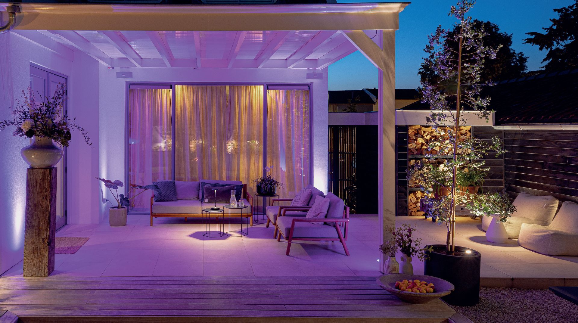 Outdoor patio at dusk, with ambient purple lighting. Includes seating area, draped curtains, and greenery.