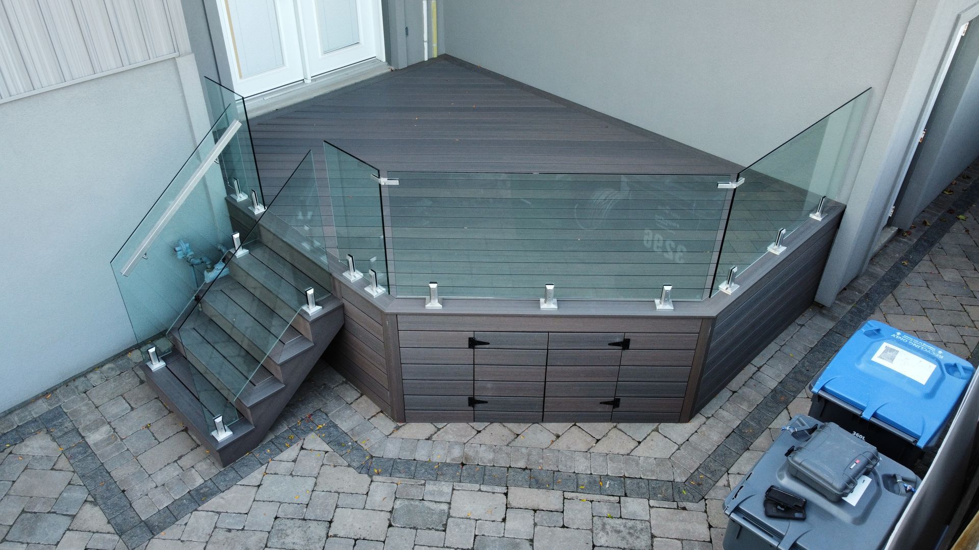 Triangular wooden deck with glass railings and steps; built-in storage, set in a paved courtyard.