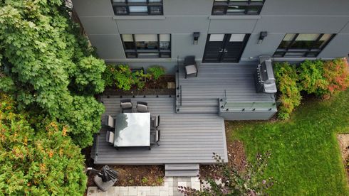 Aerial view of a gray modern house with a wooden deck, outdoor table, and green foliage.