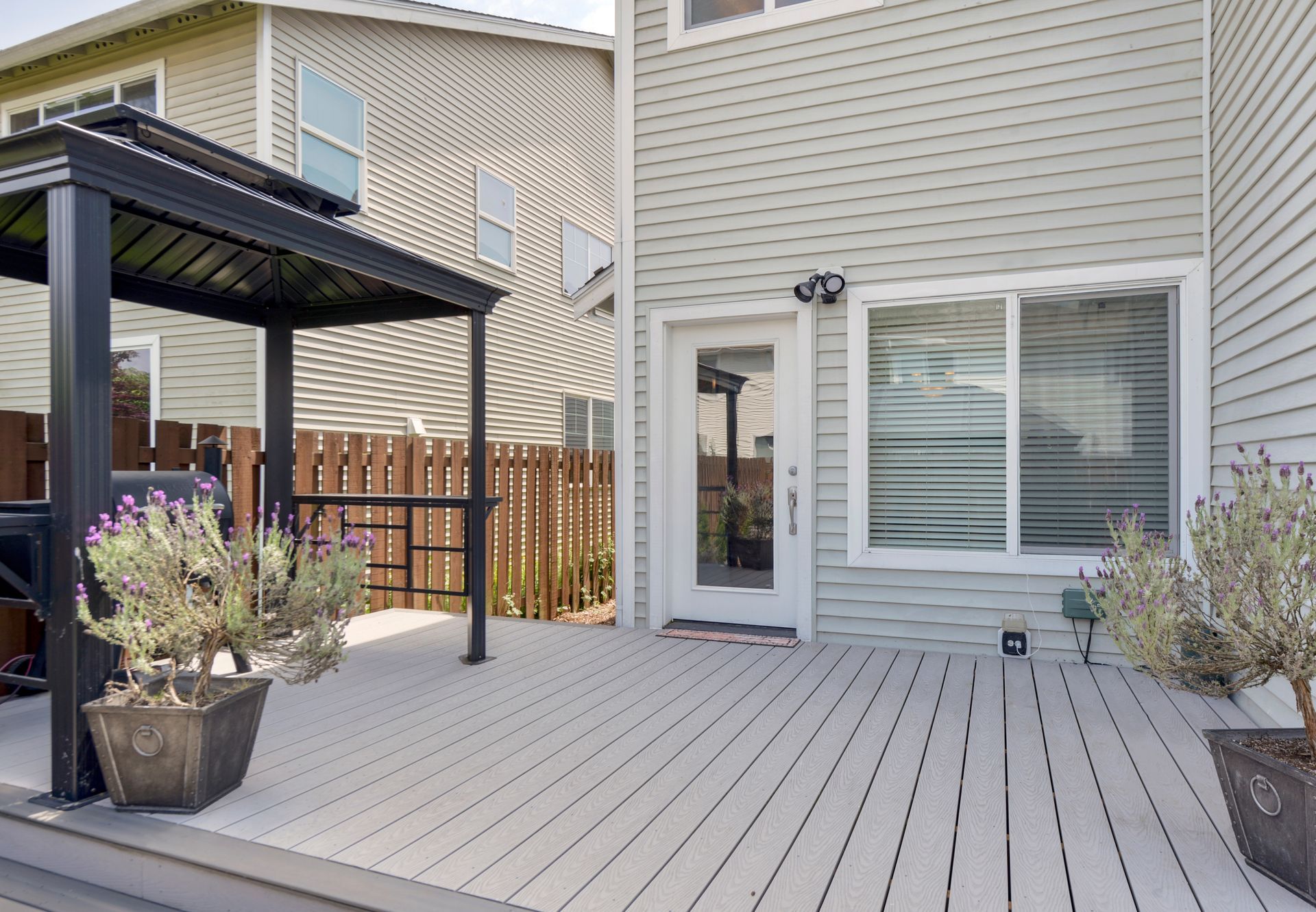 Backyard deck with gray siding, door, sliding window, black gazebo, potted plants.