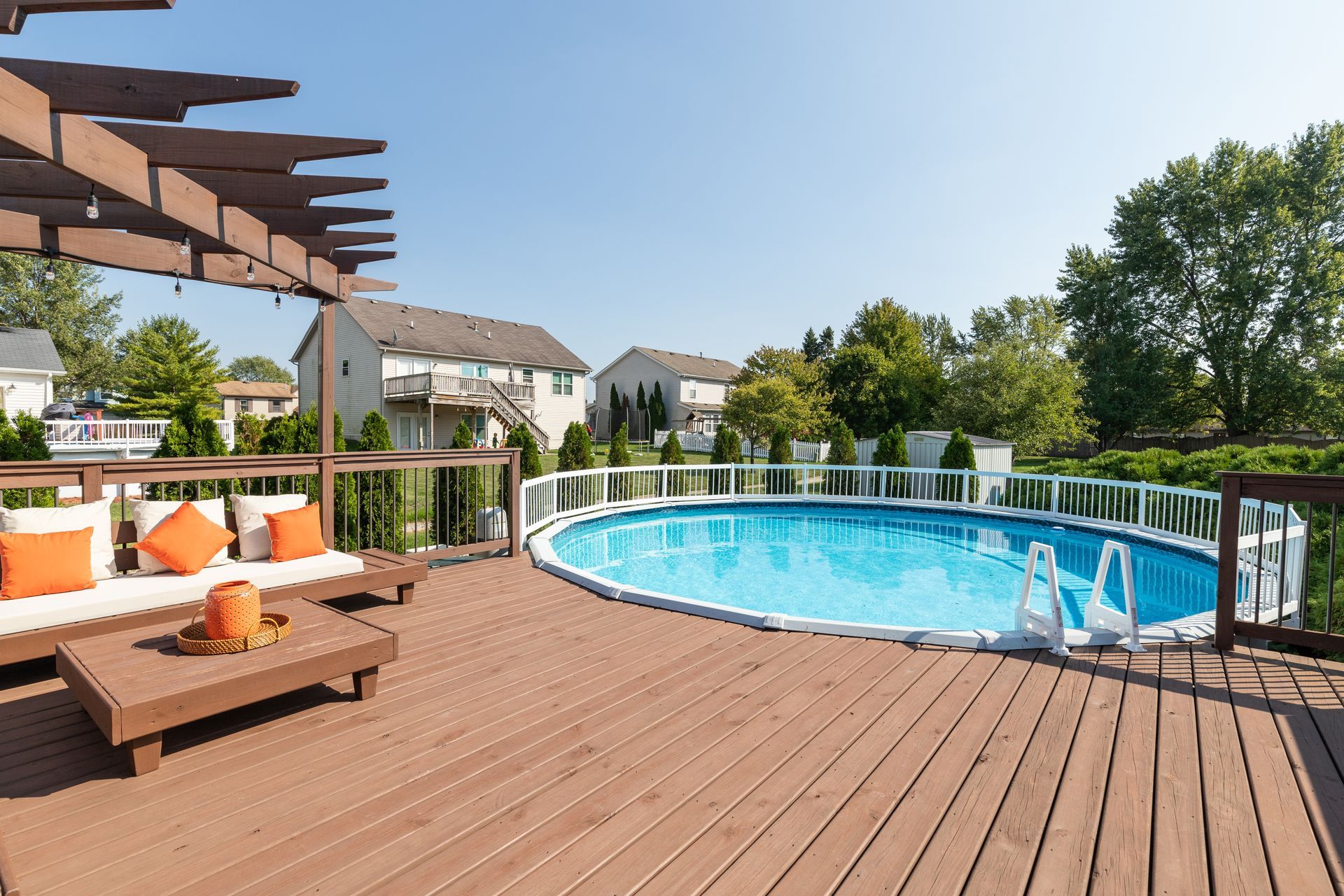 Deck with an above-ground pool. A pergola with seating and orange pillows. Houses and trees in the background.