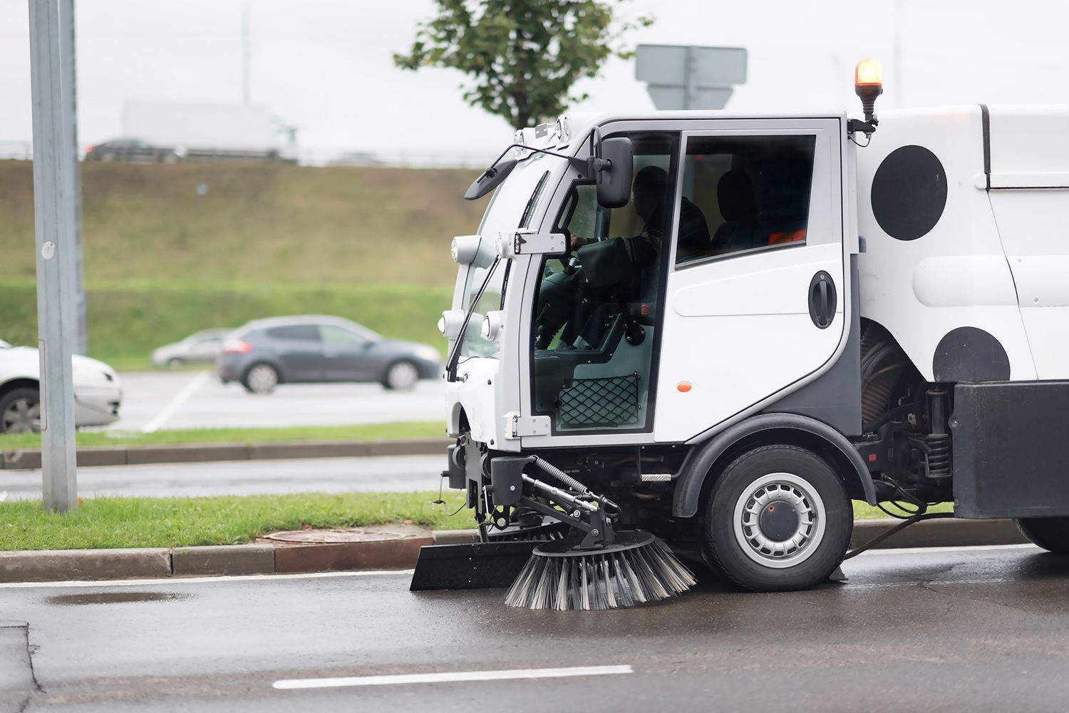 White street sweeper truck cleaning a wet road, cars in background.