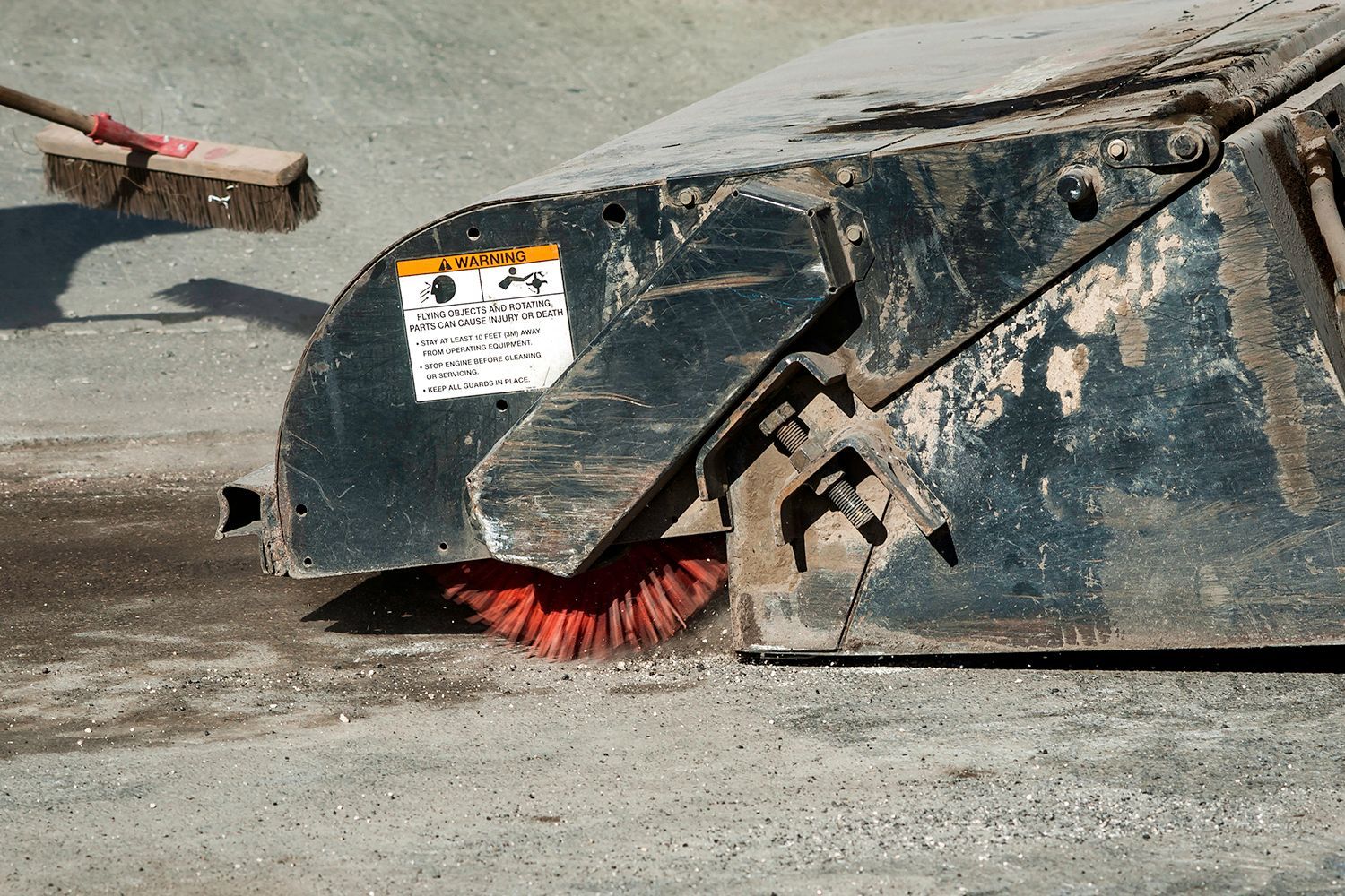 A street sweeper with a red brush cleaning a concrete surface. A broom is in the upper left. A street sweeper with a red brush cleaning a concrete surface. A broom is in the upper left.