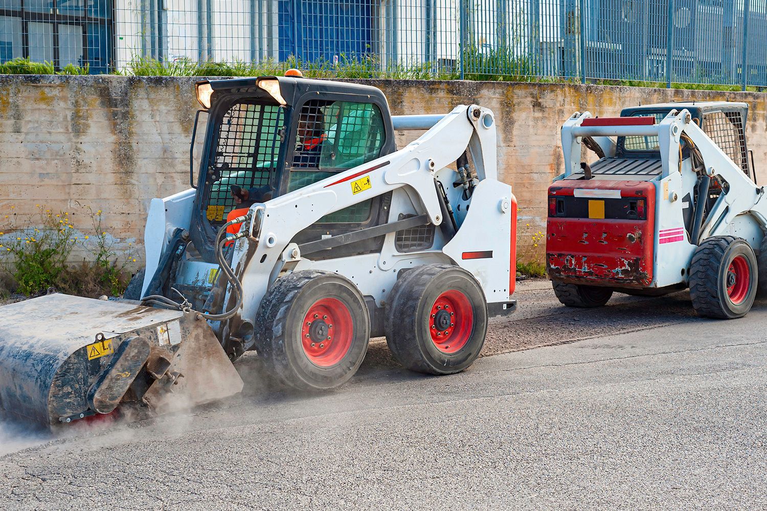 White Bobcat skid steer with a broom attachment sweeping pavement; another Bobcat nearby. White Bobcat skid steer with a broom attachment sweeping pavement; another Bobcat nearby.