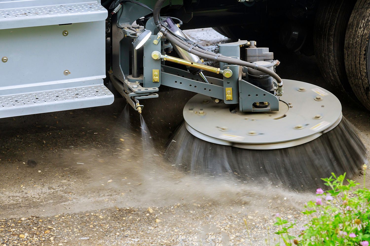 Street sweeper cleaning a gravel road, spraying water, dust rising.