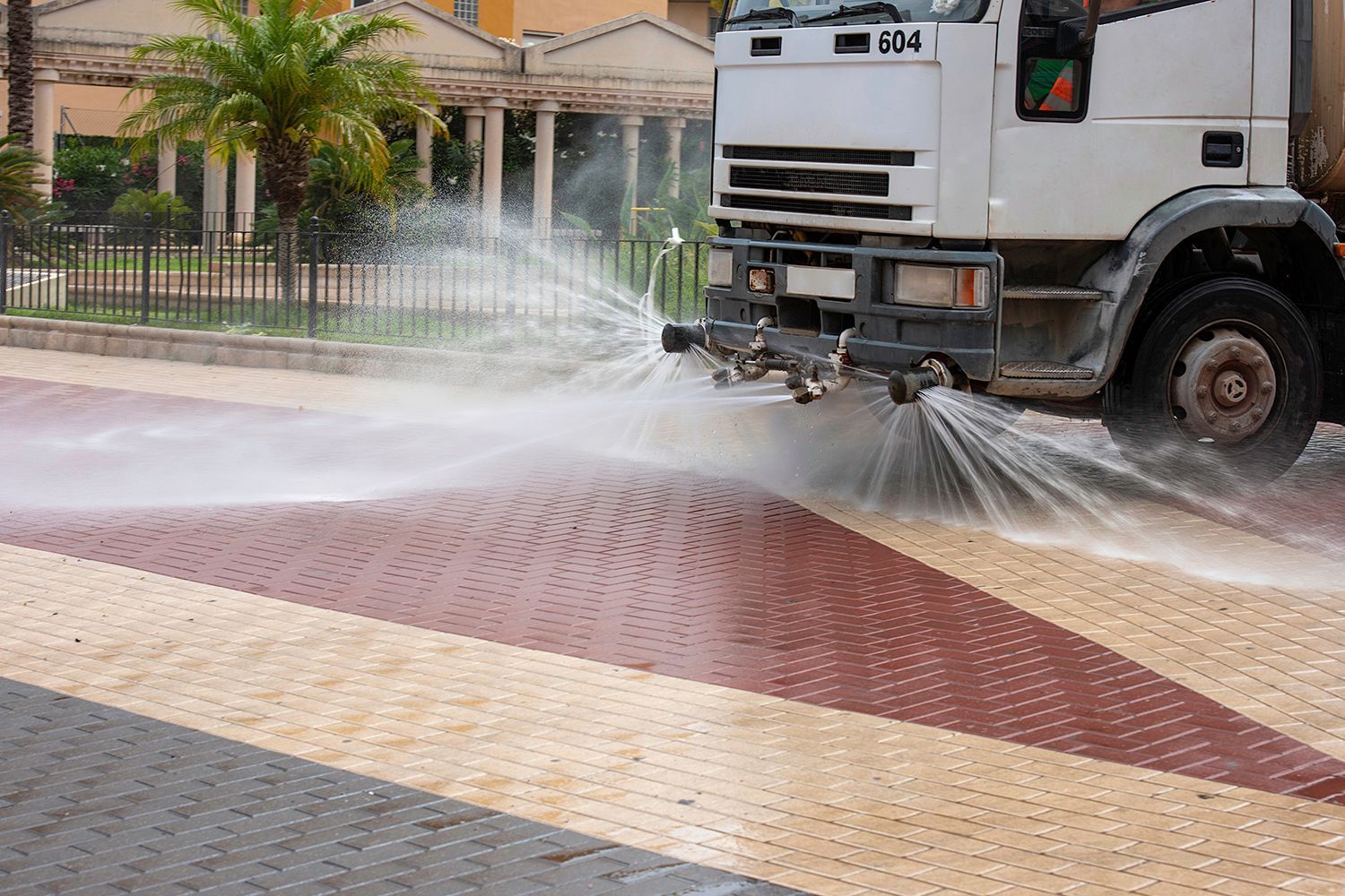 A street cleaning truck sprays water on a red and beige brick sidewalk in front of a building.