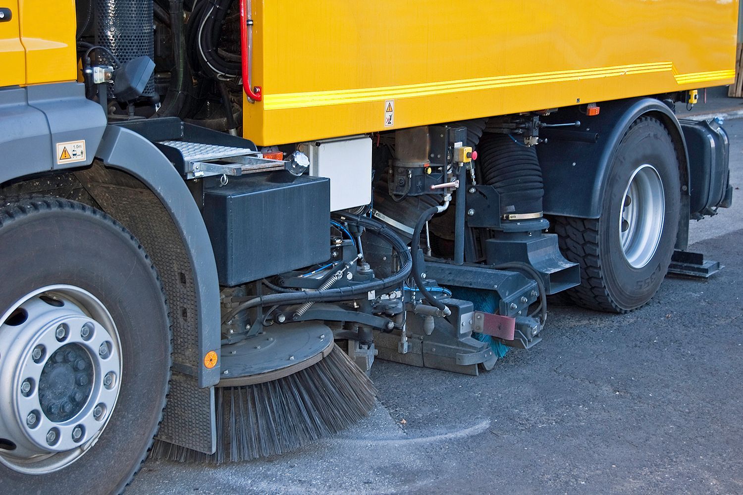 Side view of a yellow street sweeper truck with rotating brushes cleaning a paved road surface. Side view of a yellow street sweeper truck with rotating brushes cleaning a paved road surface.