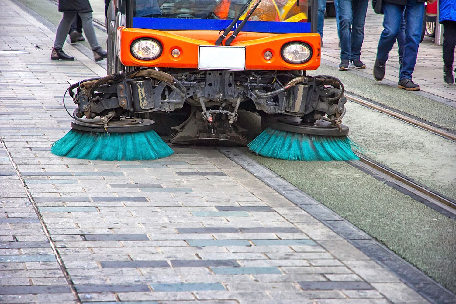 Street sweeper cleaning a brick sidewalk with green brushes, orange vehicle, and people nearby. Street sweeper cleaning a brick sidewalk with green brushes, orange vehicle, and people nearby.