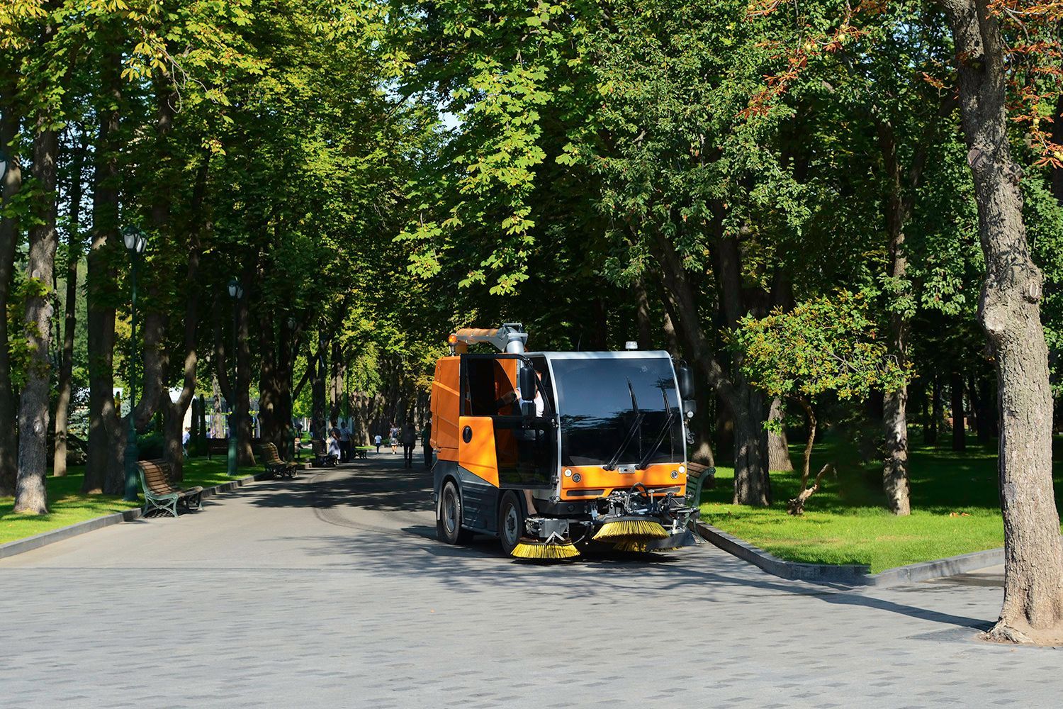 Street sweeper cleaning a tree-lined walkway in a park; orange and black vehicle, sunny day. Street sweeper cleaning a tree-lined walkway in a park; orange and black vehicle, sunny day.