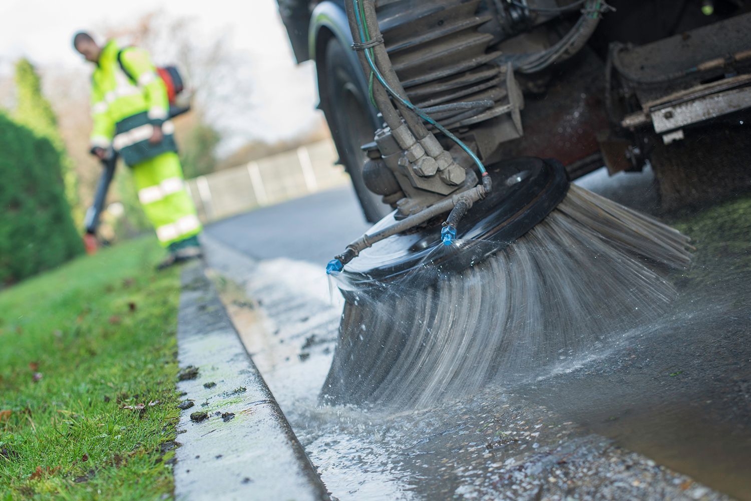 Street sweeper brushes a wet road next to a lawn; worker in reflective gear operates a leaf blower.