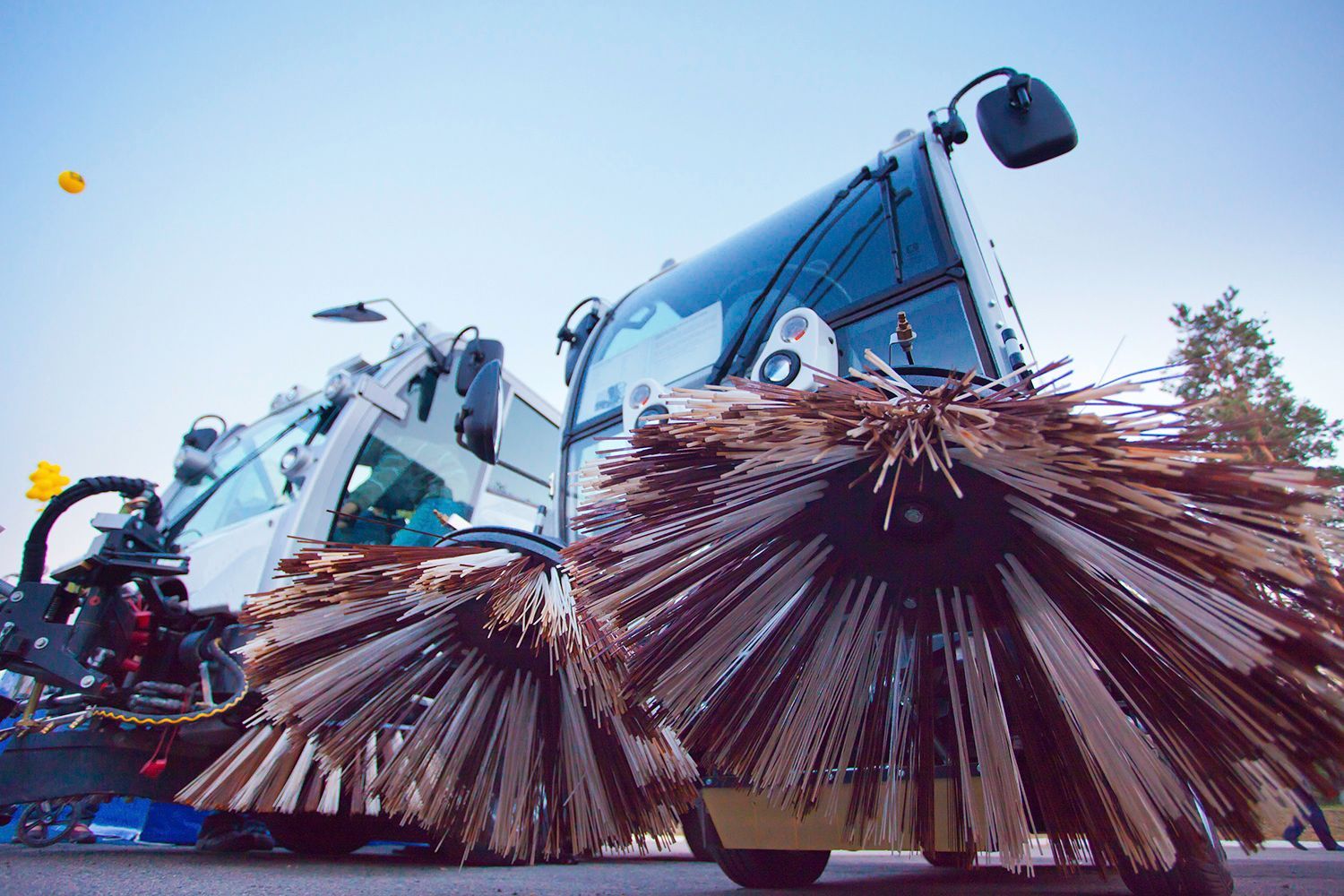 Street sweepers with large brushes, parked on a street. Blue sky in the background.