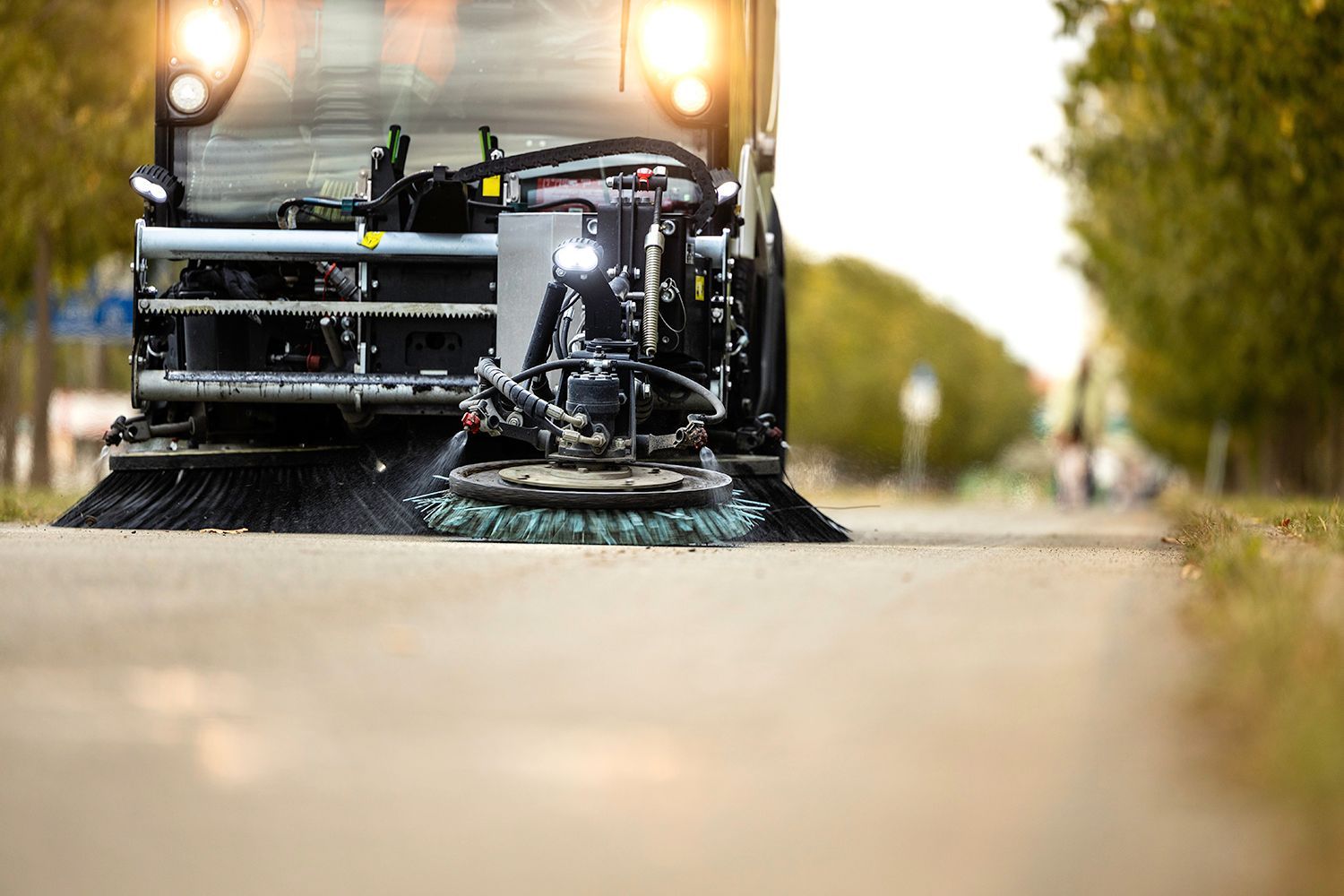 Street sweeper cleaning a road, bristles sweeping debris. Sunlight and trees in the background. Street sweeper cleaning a road, bristles sweeping debris. Sunlight and trees in the background.