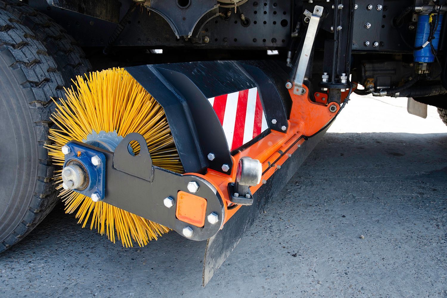 Orange and black street sweeper brush attached to a truck, sweeping a paved road.
