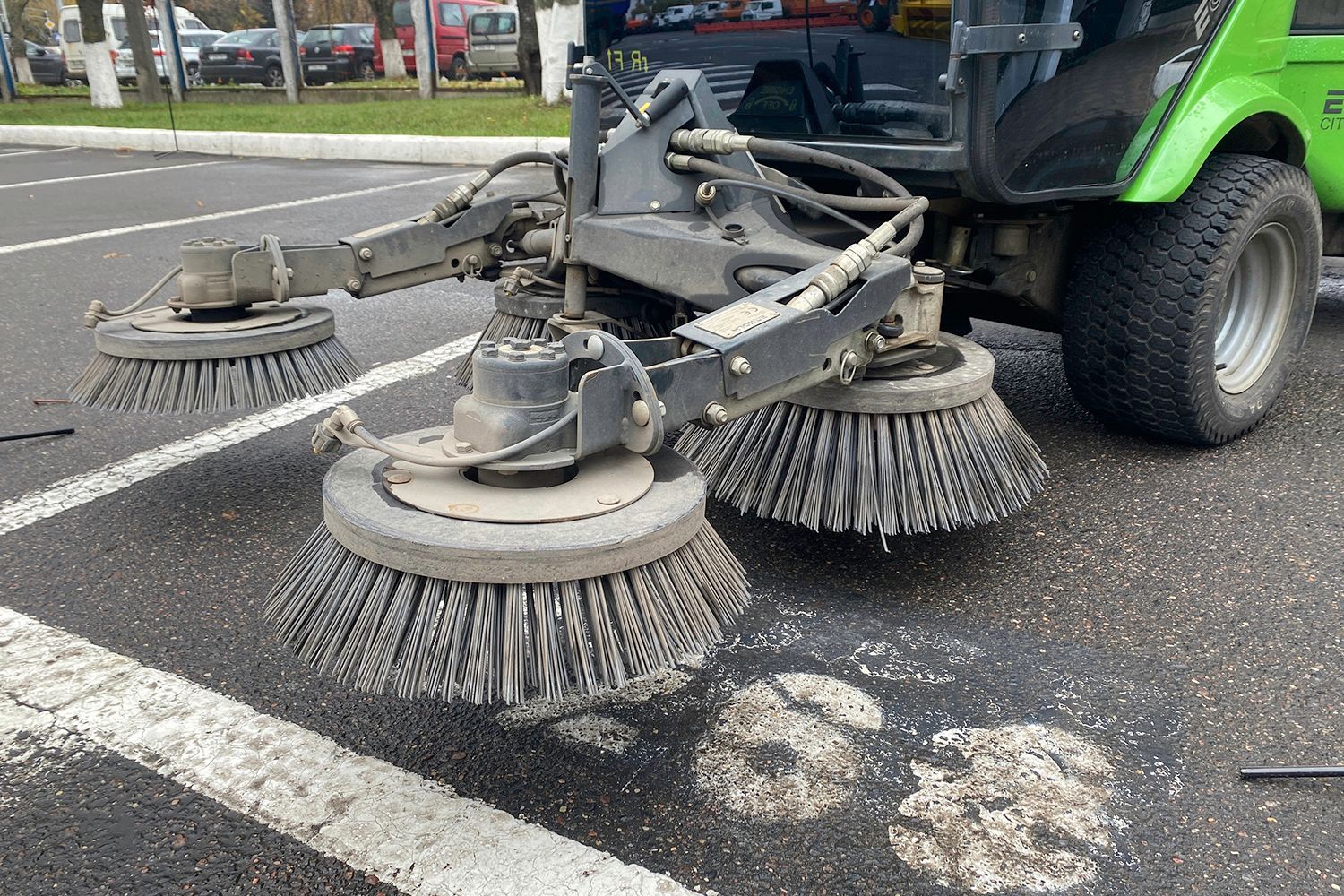 Street sweeper cleaning asphalt parking lot, featuring three rotating brushes and green vehicle.