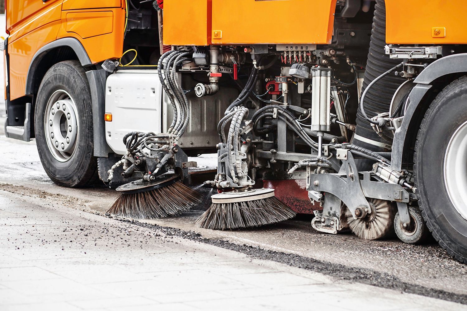 Orange street sweeper with rotating brushes cleaning a paved road.