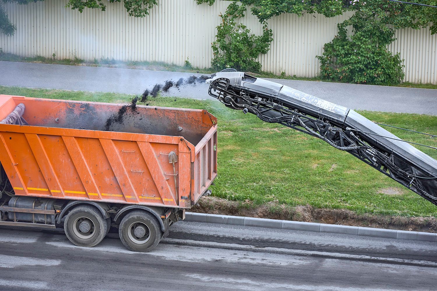 Orange dump truck being filled with asphalt by a milling machine on a street, green grass and white fence in background.