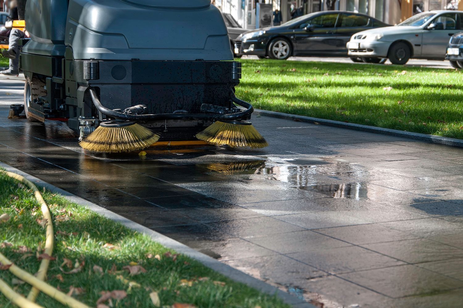 Street sweeper cleaning wet pavement beside a grassy area with parked cars in the background.