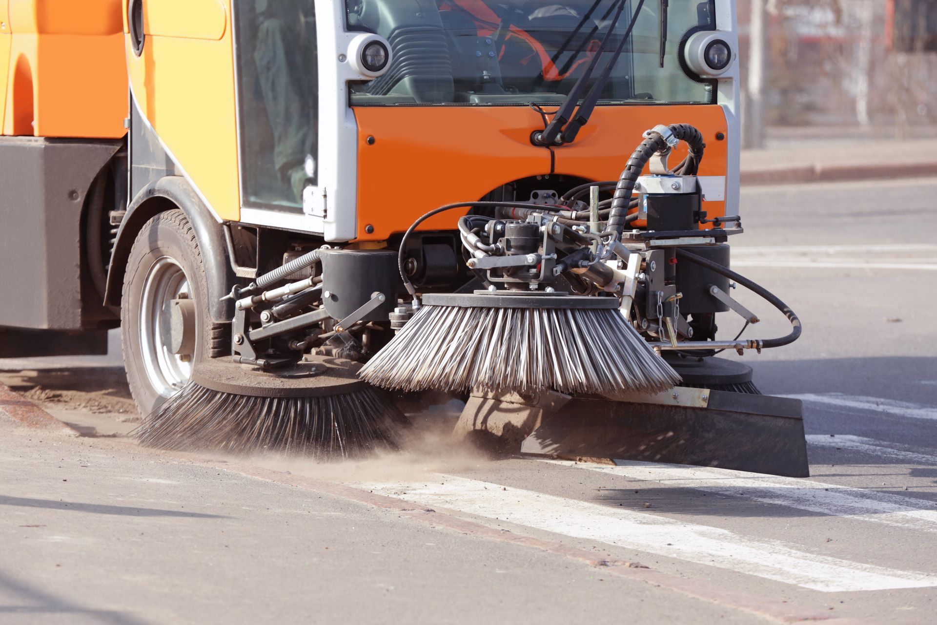 Street sweeper truck cleaning a road surface with rotating brushes and dust rising.